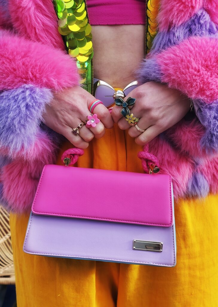 adeline wearing a fluffy pink and purple coat, green sequined jacket, bright orange trousers and holding a lilac & pink bag.