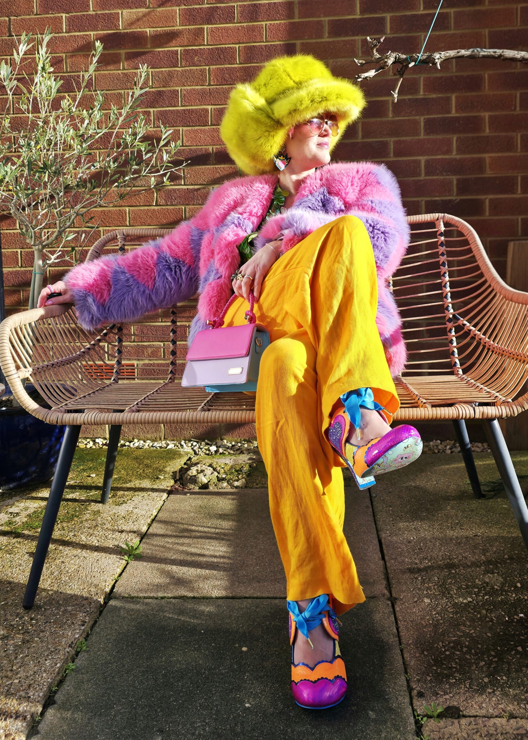 adeline wearing bright orange trousers, an oversized green beret, fluffy pink & purple hacket & bright shoes. sitting on a bench in a relaxed way.