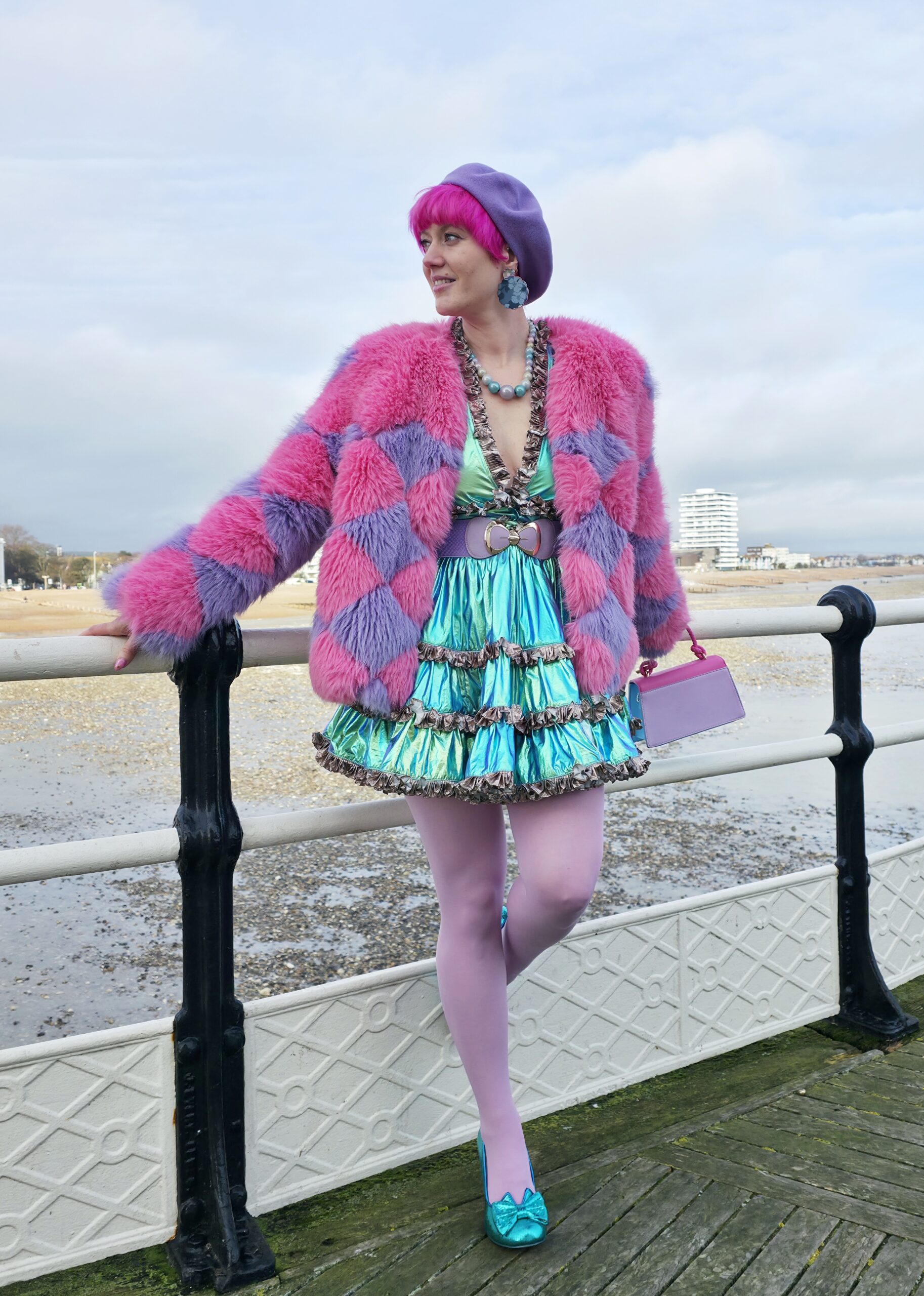 Adeline wearing a metallic teal dress, lilac tights, teal shoes, a pink & purple fluufy coat and a purple beret. Leaning against a railing by the sea