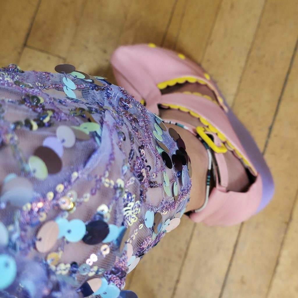 A top down photo of Adeline's leg. She is wearing a sparkly lilac dress, and a pair of pink and lilac shoes with soft yellow scalloped trims.